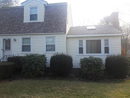 A small, white, two-story house with a shingled roof and a chimney. Bushes and hedges line the front, under a clear, bright sky.