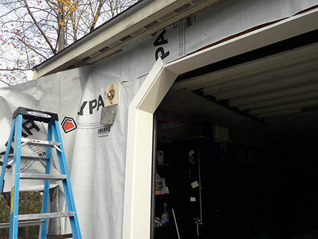 Partially constructed building exterior with visible insulation wrap, a blue ladder, and an open garage.