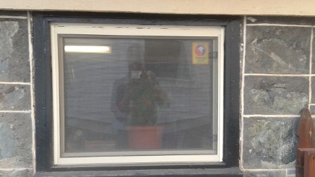A window is set in a stone wall, with a faint reflection of a person holding a camera and a potted plant inside.