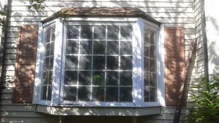 A sunlit bay window with grid panes is framed by wooden shutters on a beige house.