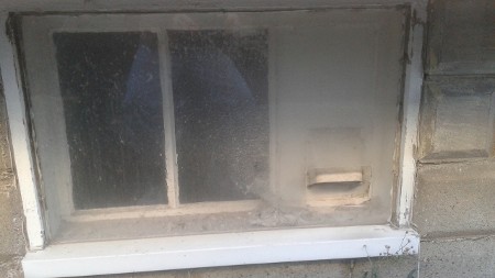 A small, dusty, and dirty basement window with peeling paint and cobwebs.