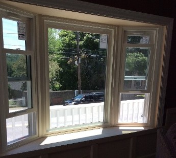 A sunlit bay window with three sections, partially open, frames a view of a suburban street, greenery, and a parked black car, creating a serene scene.