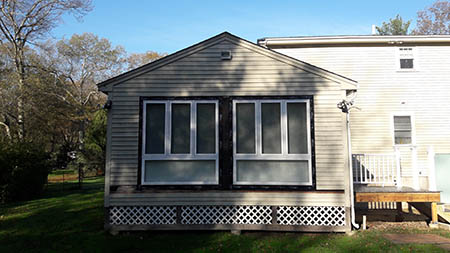 A small house extension with beige siding and two large windows, set in a grassy yard with trees in the background under a clear blue sky.