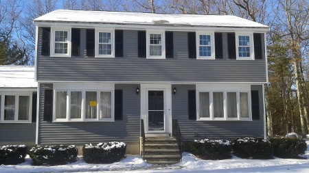 Two-story grey house with black shutters, snow-covered roof, and surrounding bushes.