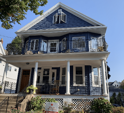 Two-story blue house with white trim, featuring a balcony and columns, set against a clear sky.